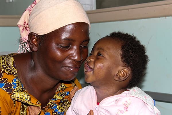 A woman with a bright orange dress is leaning into her daughter who has a cleft palate and who is smiling.