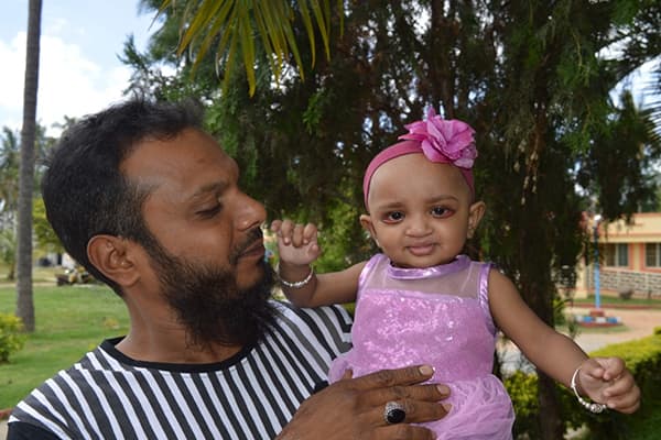 A man is hugging and holding his baby daughter who is wearing a pink dress under a palm tree.