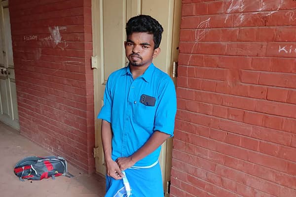 A young man with black hair and a moustache is wearing a blue collard shirt and standing in front of a red brick wall