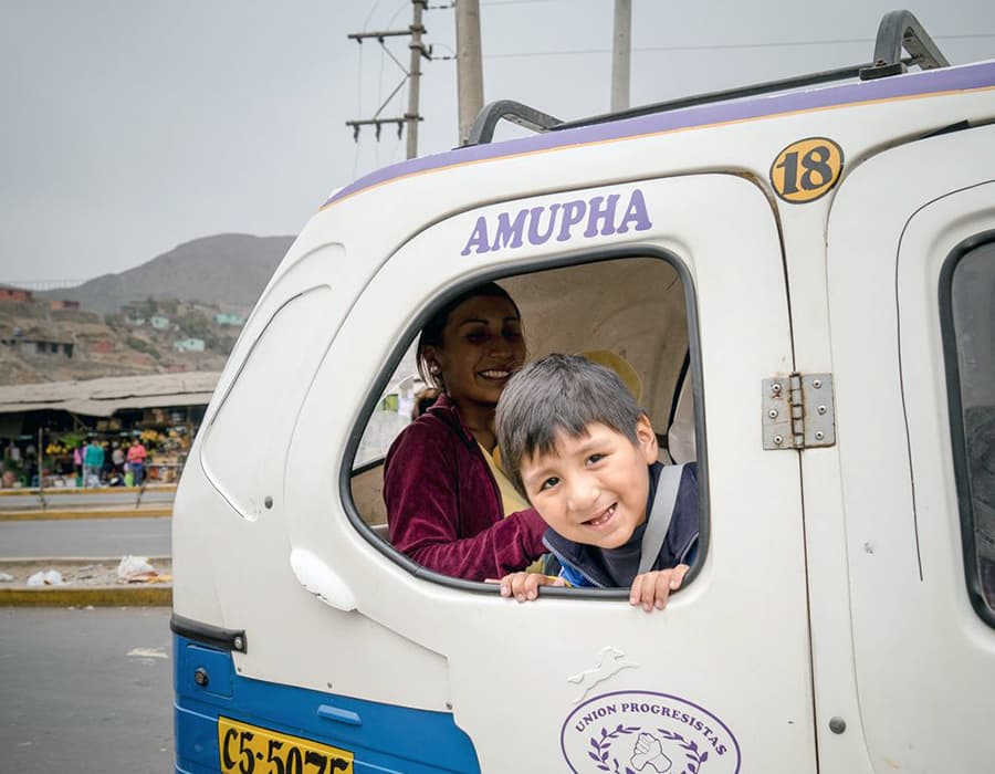 A smiling child leaning out of a van window with his mother smiling in the background