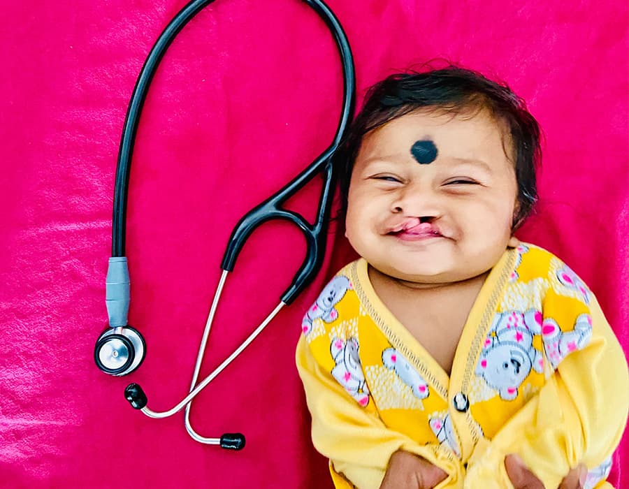 A child with cleft lip laying down beside a stethoscope on a pink background